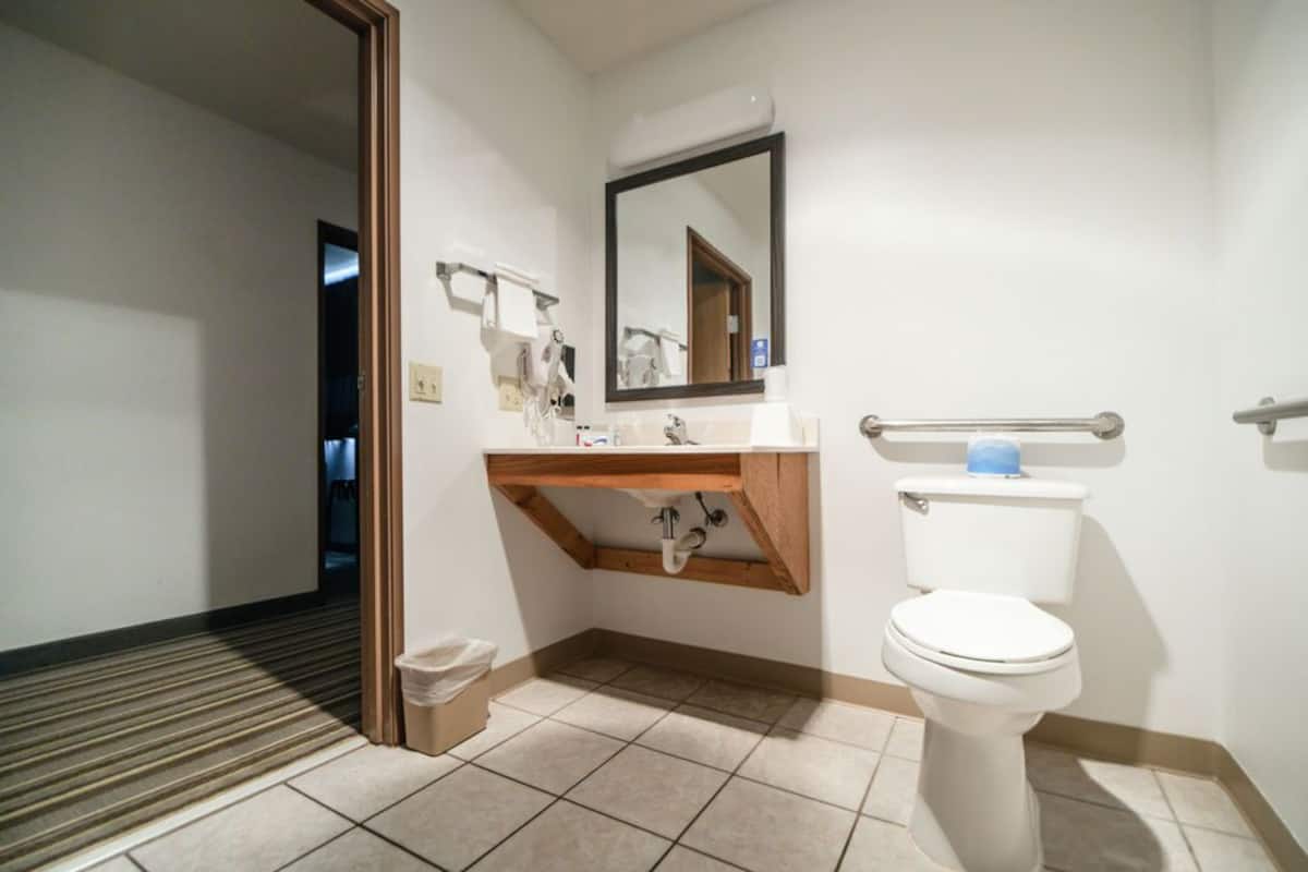 Een badkamer met een wit wastafel, spiegel en toilet. Er is een houten plank onder de wastafel en een prullenbak aan de linkerkant.