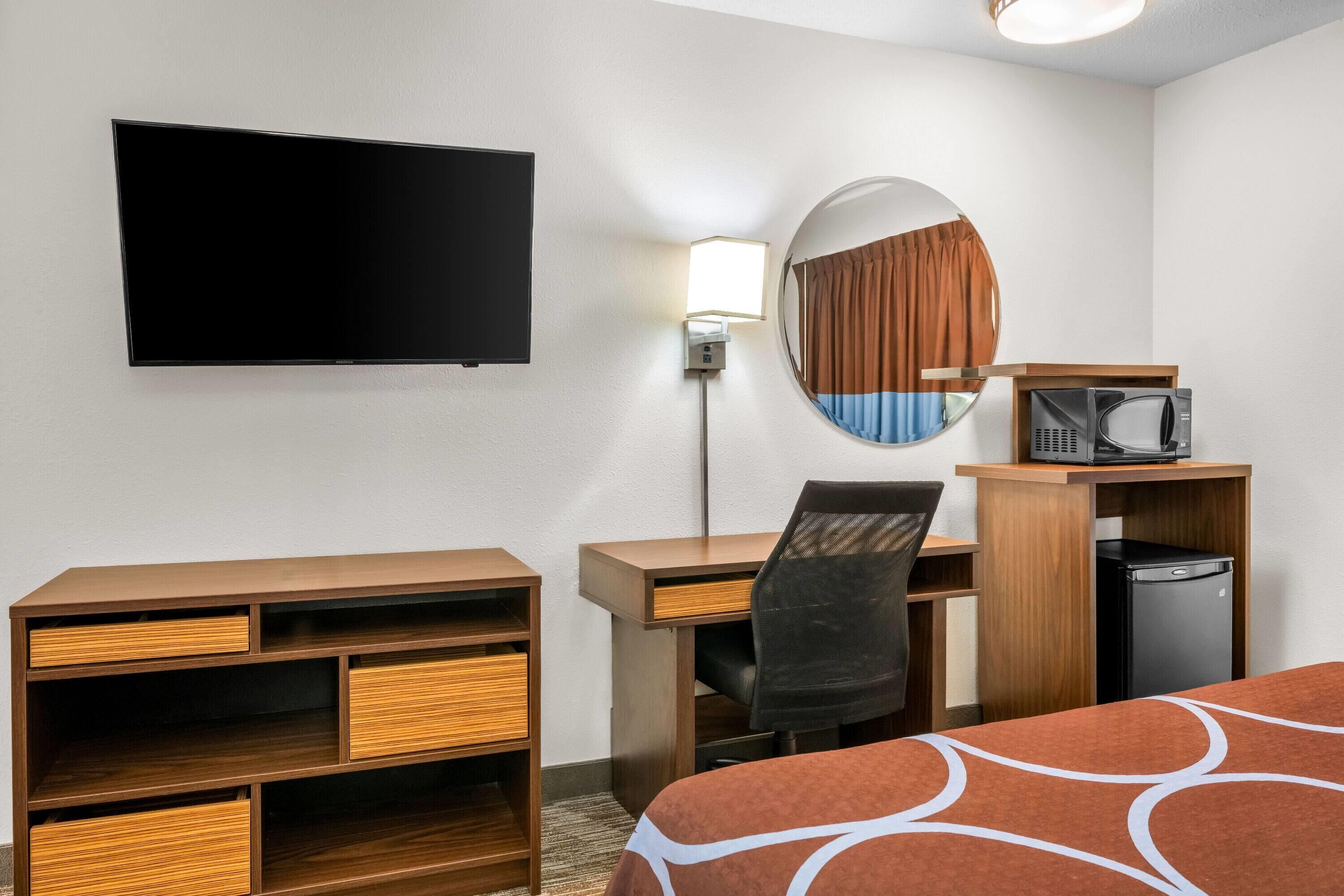 A hotel room with a bed, a desk with a chair, a television, a mirror, a lamp, a microwave, and a small refrigerator.