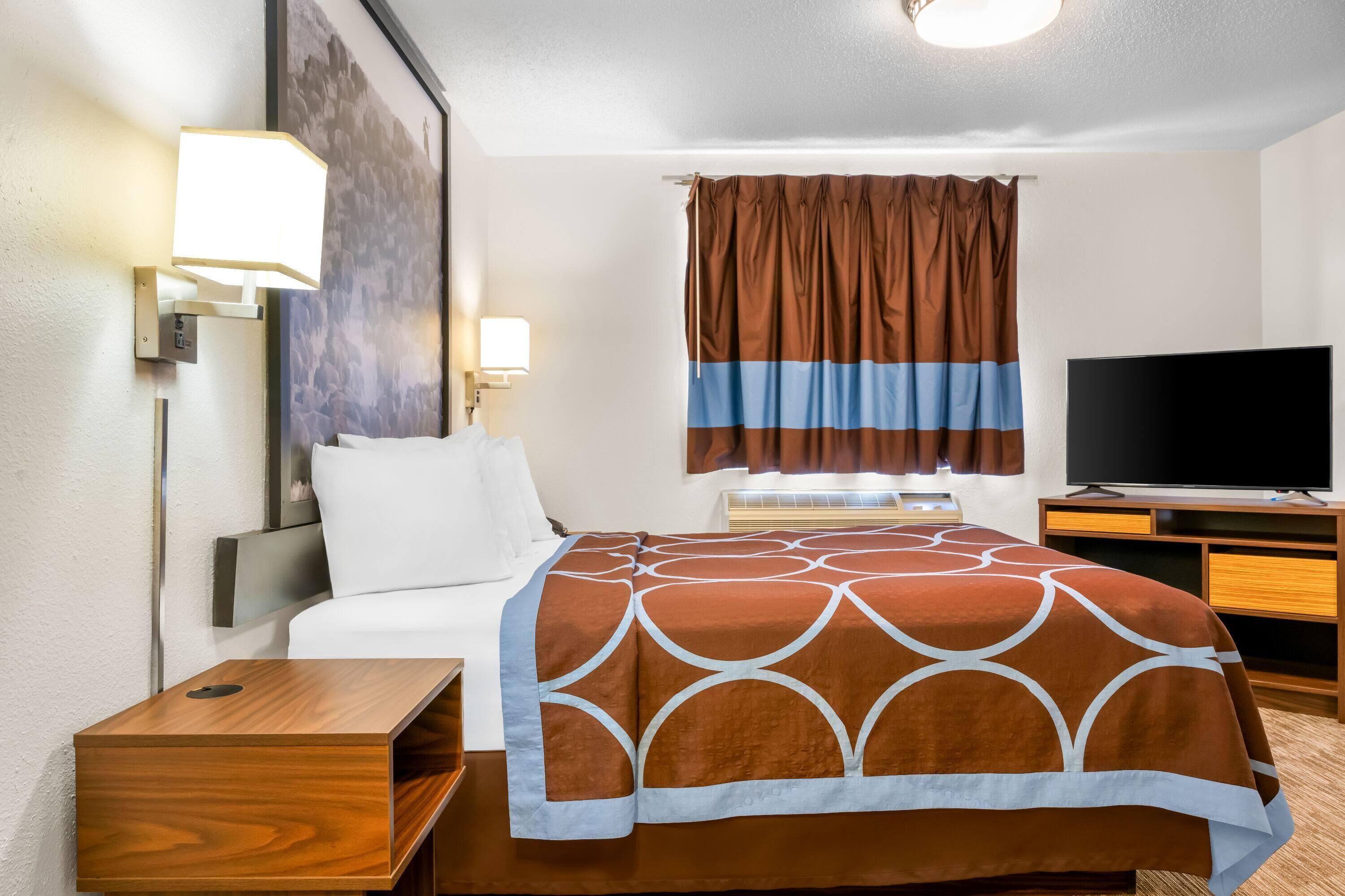 A hotel room with a bed, a TV, a desk, and a wall-mounted lamp.