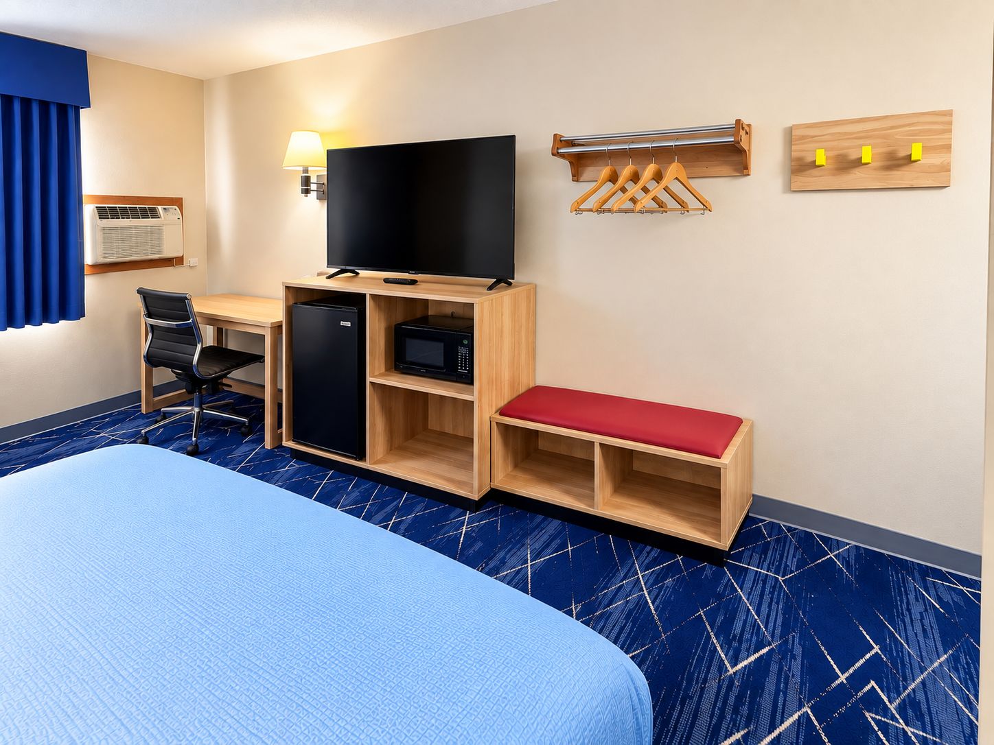 A hotel room with a bed, desk, chair, refrigerator, microwave, television, and a wall-mounted coat rack.