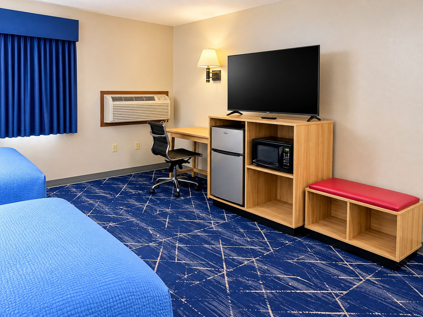 A hotel room with a bed, a desk with a computer, a refrigerator, a microwave, a television, and a red bench.