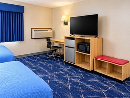 Habitación de hotel con una cama, un escritorio con computadora, refrigerador, microondas, televisor y un banco rojo.