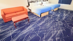 Hotellihuone, jossa on sininen matto, kaksi sänkyä valkoisilla vuodevaatteilla, punainen sohva ja pieni pöytä.