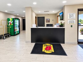 Zona de recepción de hotel con refrigerador de 7UP, mostrador de recepción y letrero de vigilancia las 24 horas.
