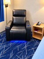 Una silla de masaje negra con panel de control, una mesita de noche con teléfono y una alfombra azul estampada.