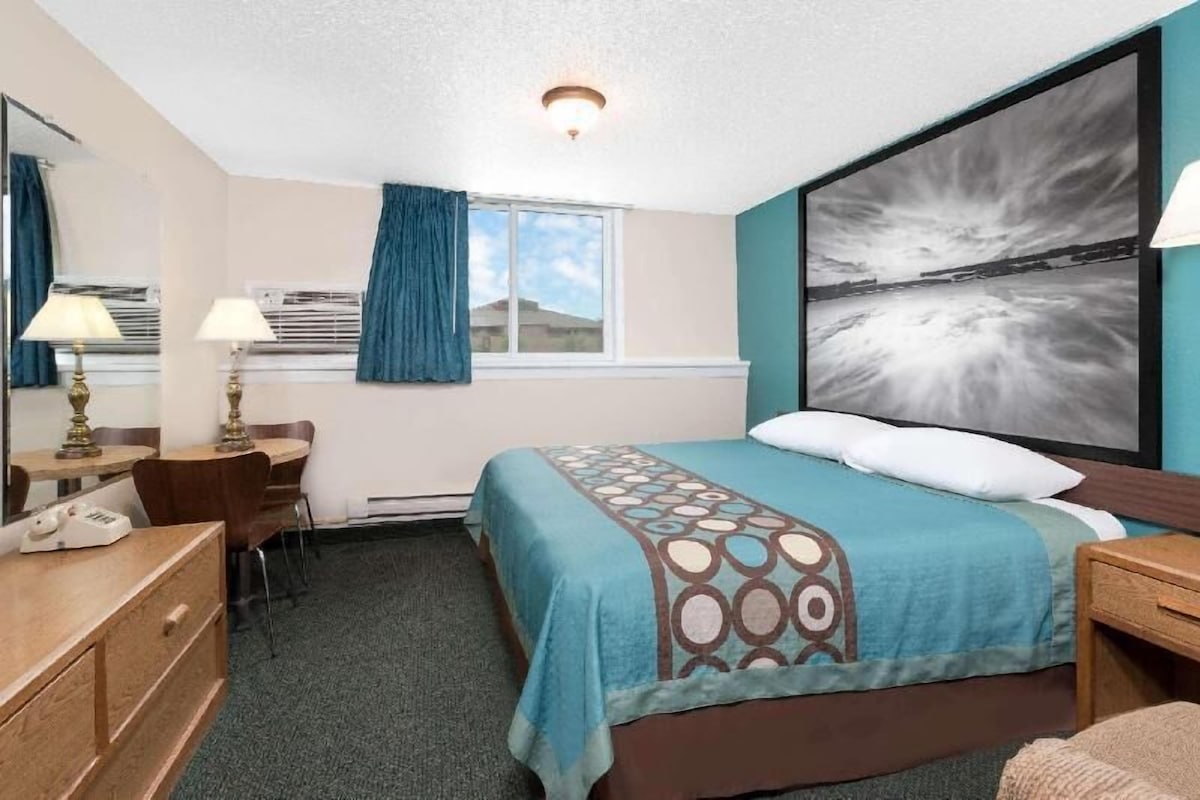 Habitación de hotel con cama, escritorio, silla, cómoda, ventana con cortinas y un cuadro abstracto grande en la pared.