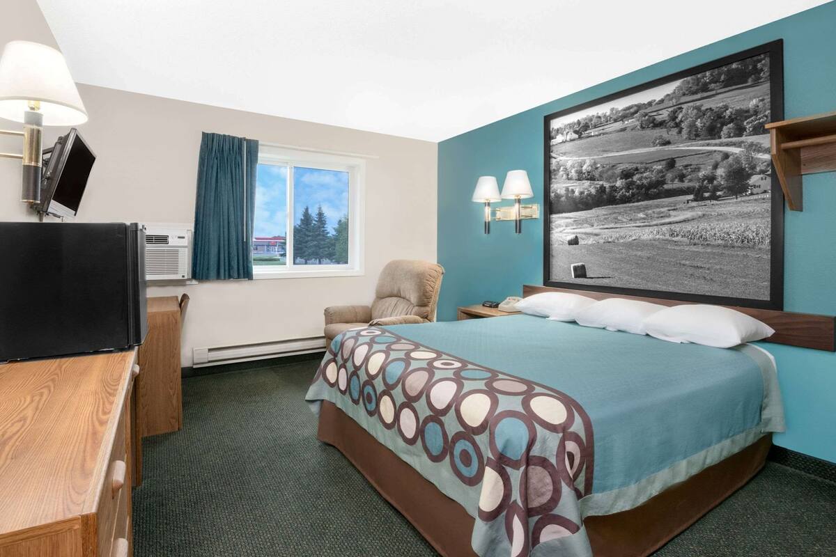 A hotel room with a bed, a desk, a chair, a TV, a window with curtains, and a large landscape painting on the wall.
