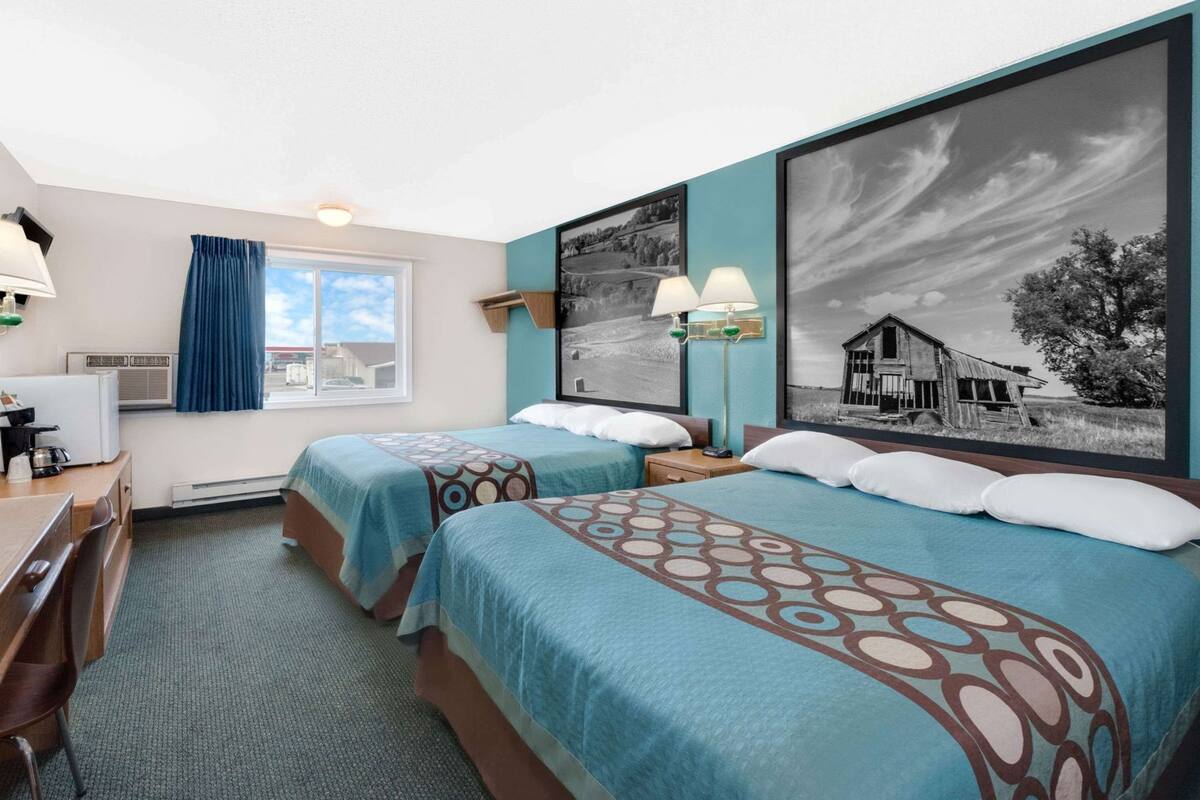 A hotel room with two beds, a desk, a chair, a lamp, and large framed pictures on the wall.