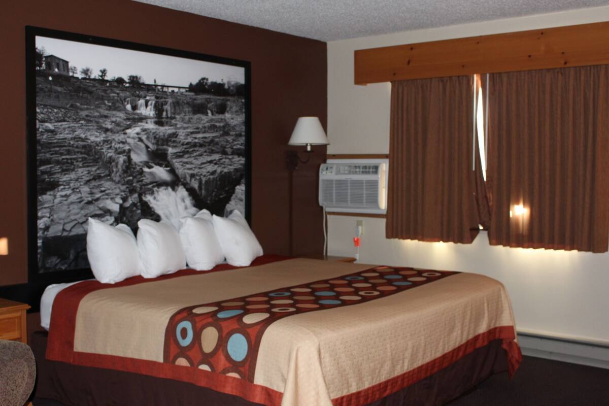 A hotel room with a large bed, a wall-mounted air conditioner, a window with curtains, and a framed photograph on the wall.