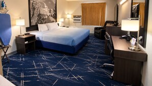 A hotel room with a large bed, a desk with a chair, a desk lamp, a chair, a wall-mounted TV, and a large painting.