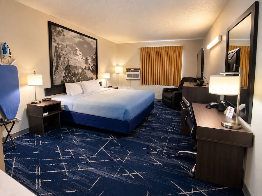 A hotel room with a large bed, a desk with a chair, a desk lamp, a chair, a wall-mounted TV, and a large painting.