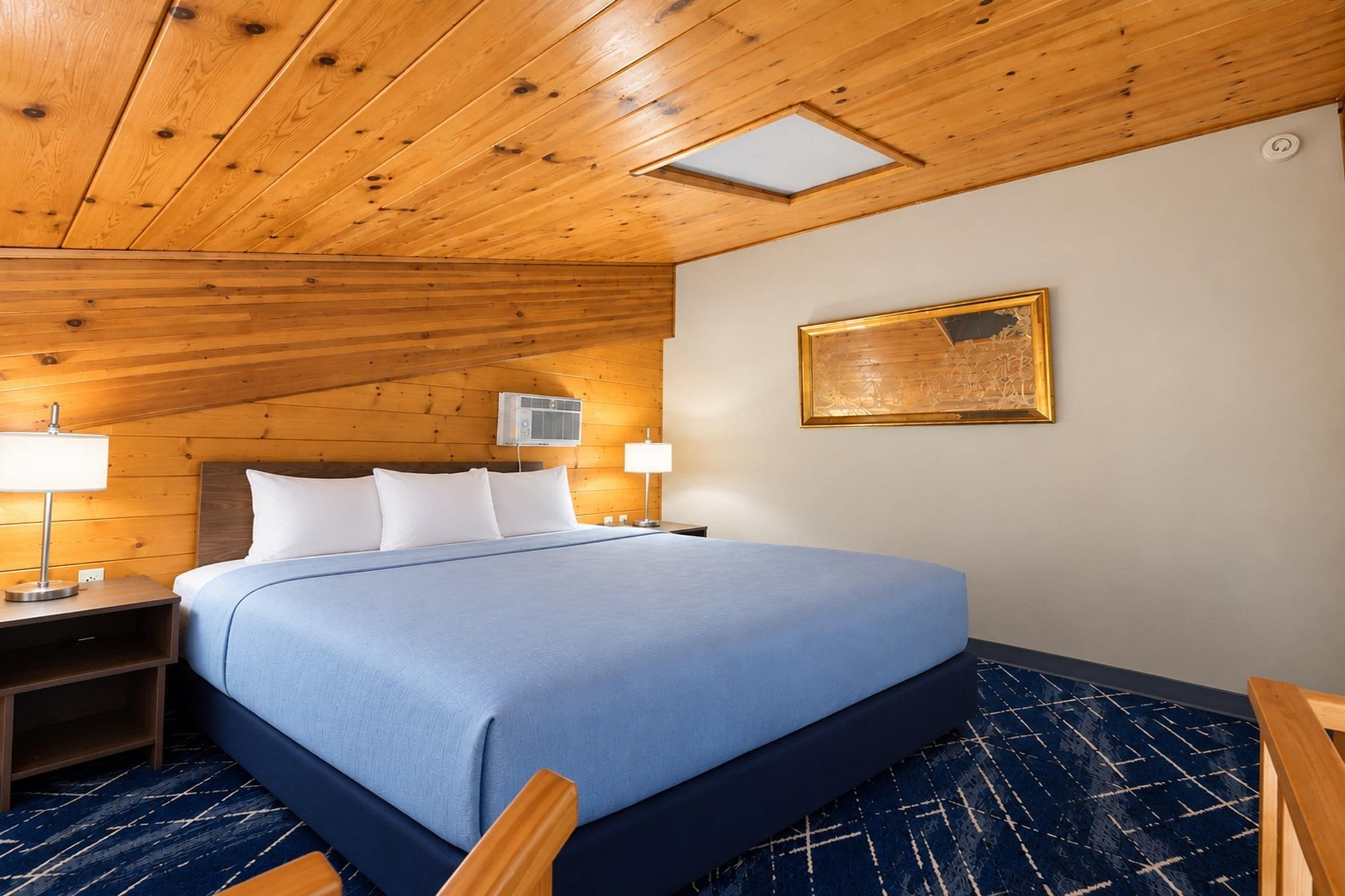Een hotelkamer met een houten plafond, een bed met een blauwe sprei, twee nachtlampjes en een wandgemonteerde airconditioning.