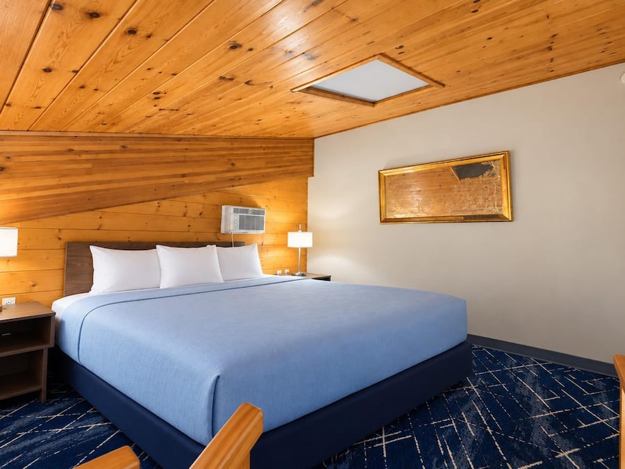 A hotel room with a wooden ceiling, a bed with a blue bedspread, two bedside lamps, and a wall-mounted air conditioning unit.