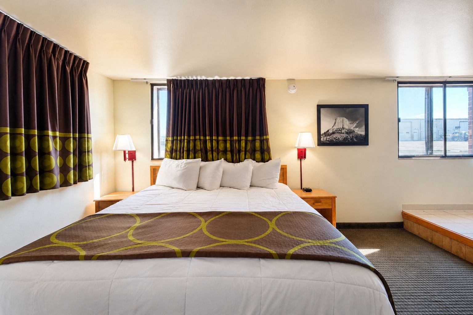 Quarto de hotel com cama, duas luminárias de cabeceira, janela com cortinas e um quadro pendurado na parede.