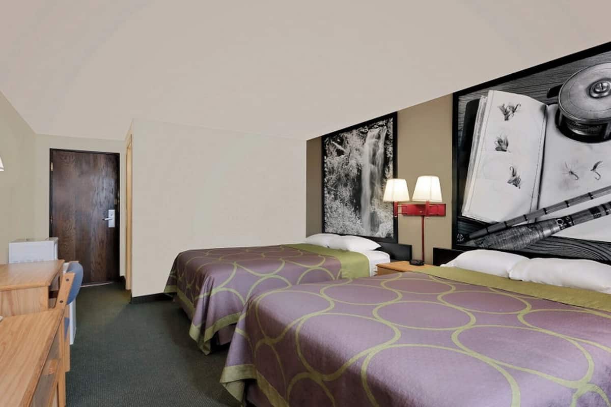 A hotel room with two beds, a desk, a chair, a nightstand, a lamp, and a large painting on the wall.