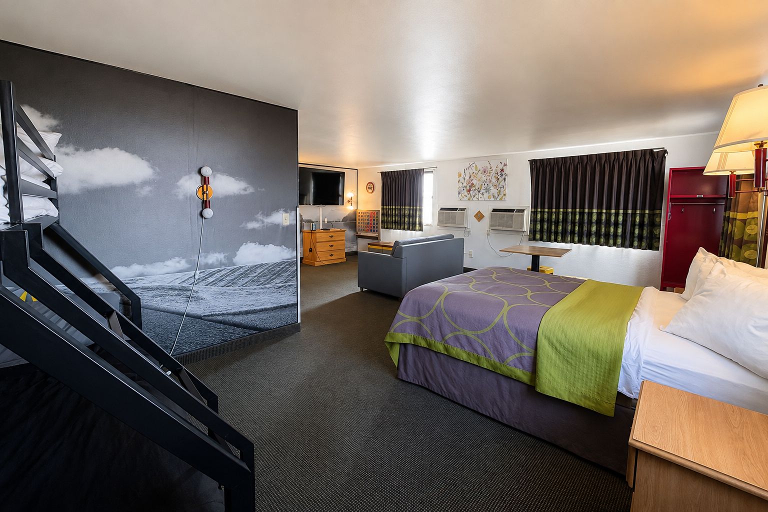 A hotel room with a bed, a desk, a chair, a dresser, and a wall with a mural.