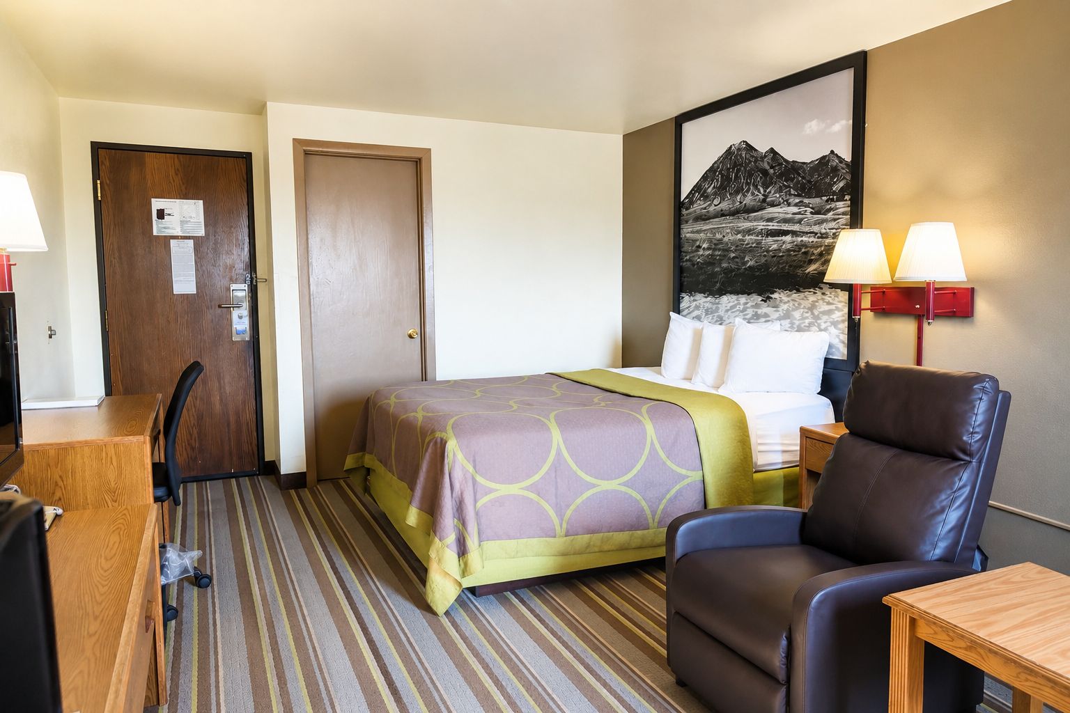 Habitación de hotel con cama, escritorio, silla, lámpara y un cuadro en la pared.