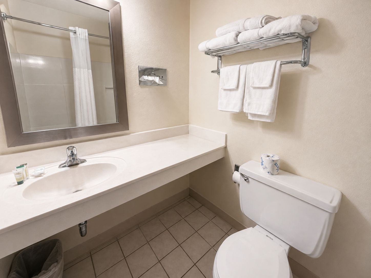 A bathroom with a white sink, mirror, and toilet. There is a towel rack with folded towels above the toilet.