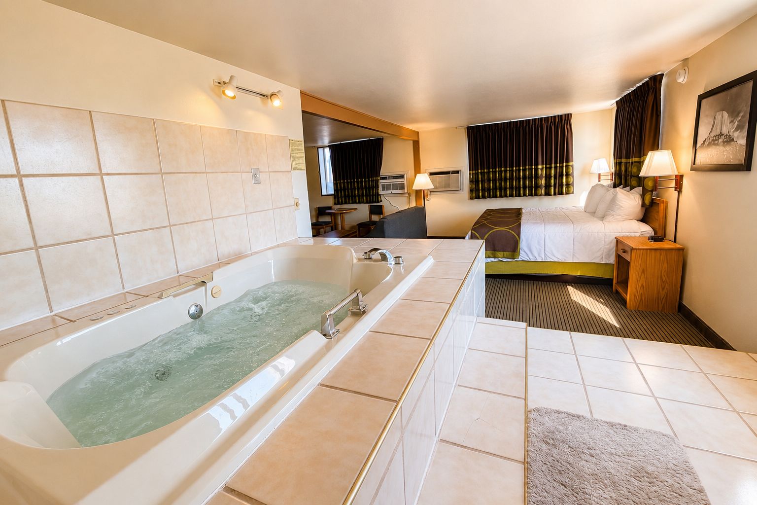 A hotel room with a large bathtub, a bed, a bedside table with a lamp, and a picture on the wall.