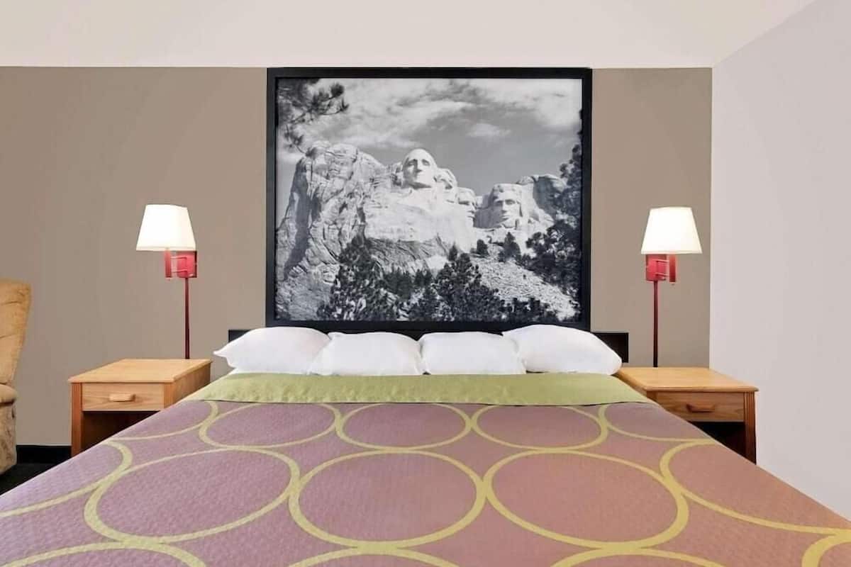 A hotel room with a bed, two bedside tables, two lamps, a large framed picture of a mountain, and a green throw blanket.