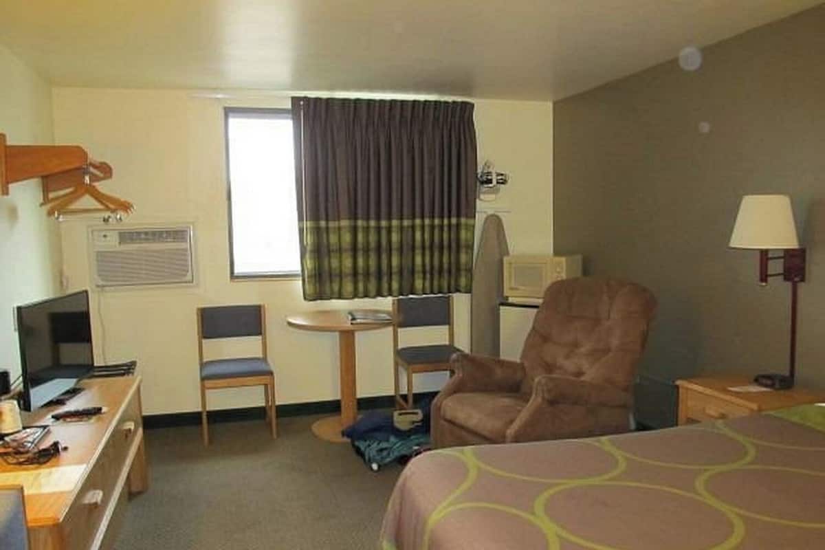 A hotel room with a bed, a desk with a computer, two chairs, a TV, a lamp, and a window with curtains.