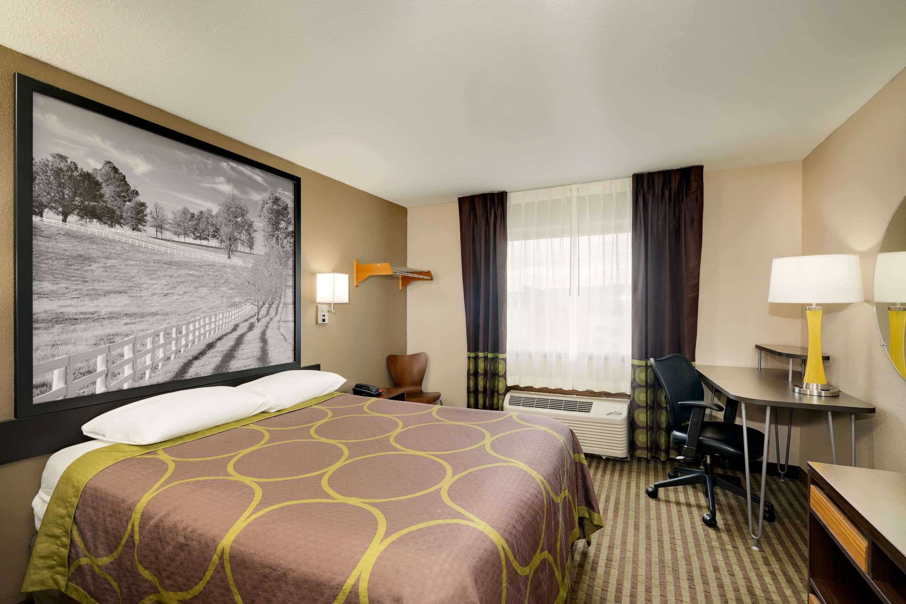 A hotel room with a large bed, a desk, a chair, a desk lamp, a wall-mounted lamp, a window with curtains, and a framed picture on the wall.