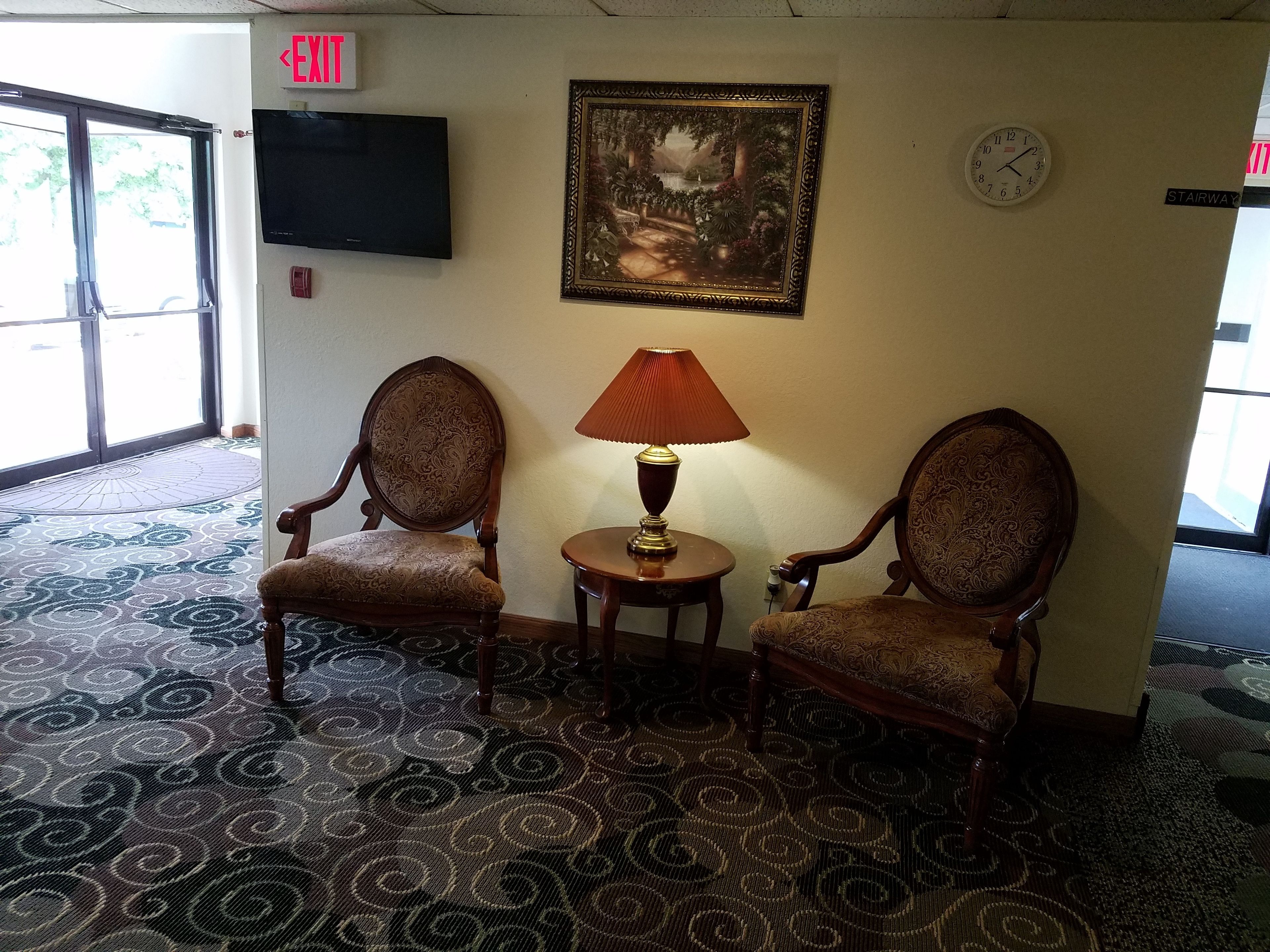 A lobby with two ornate chairs, a table with a lamp, a framed picture, a clock, and an exit sign.
