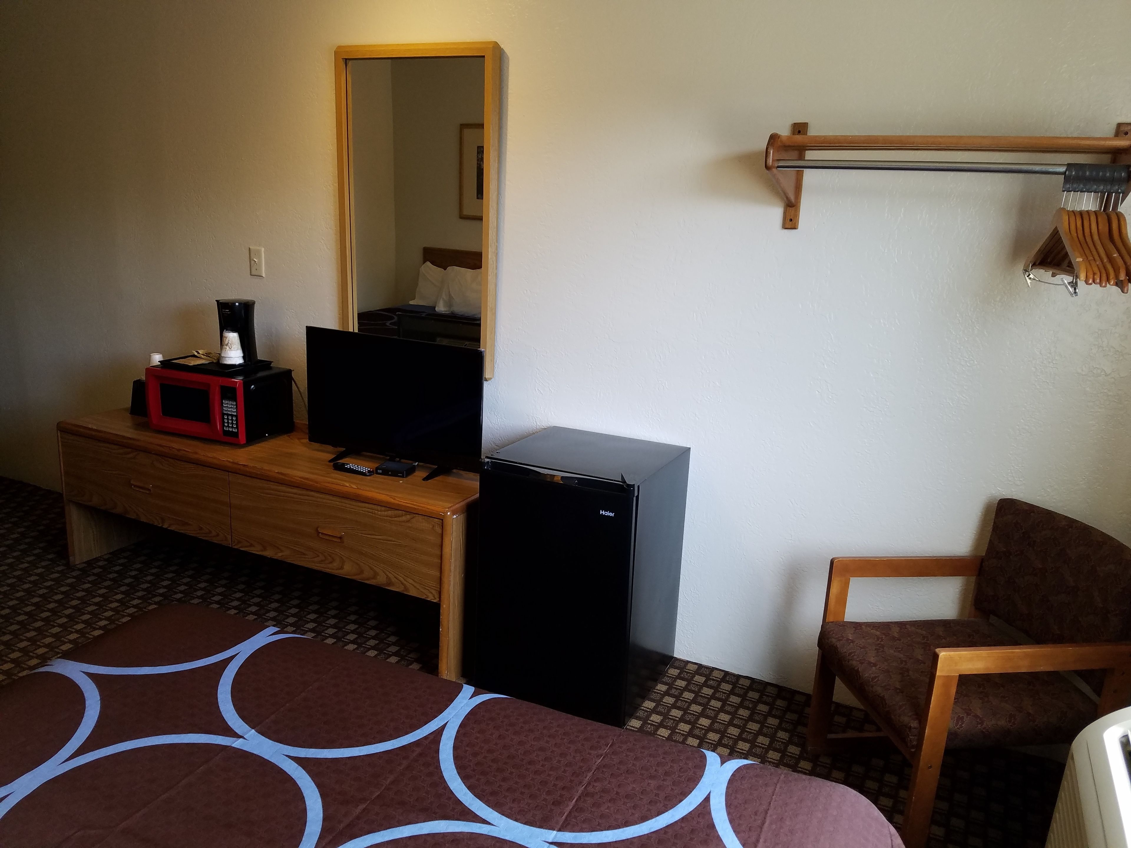A hotel room with a wooden desk, a television, a small refrigerator, a chair, and a bed with a patterned bedspread.