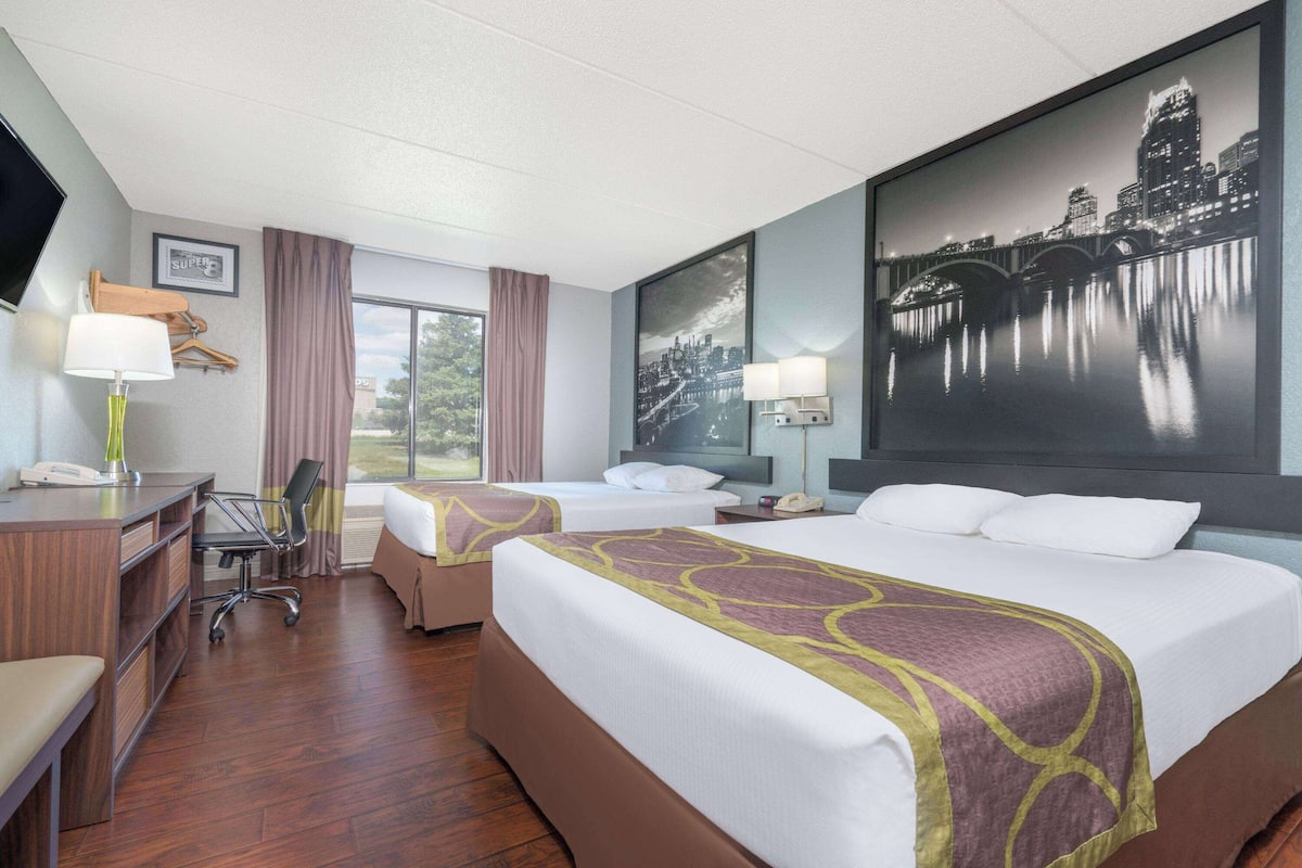 A hotel room with two beds, a desk, a chair, a TV, and a large cityscape painting.