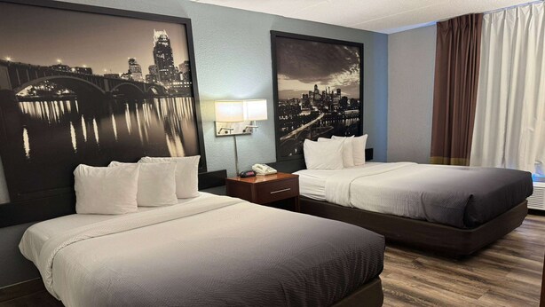 Habitación de hotel con dos camas, una mesita de noche, una lámpara y dos grandes cuadros con vistas urbanas en las paredes.