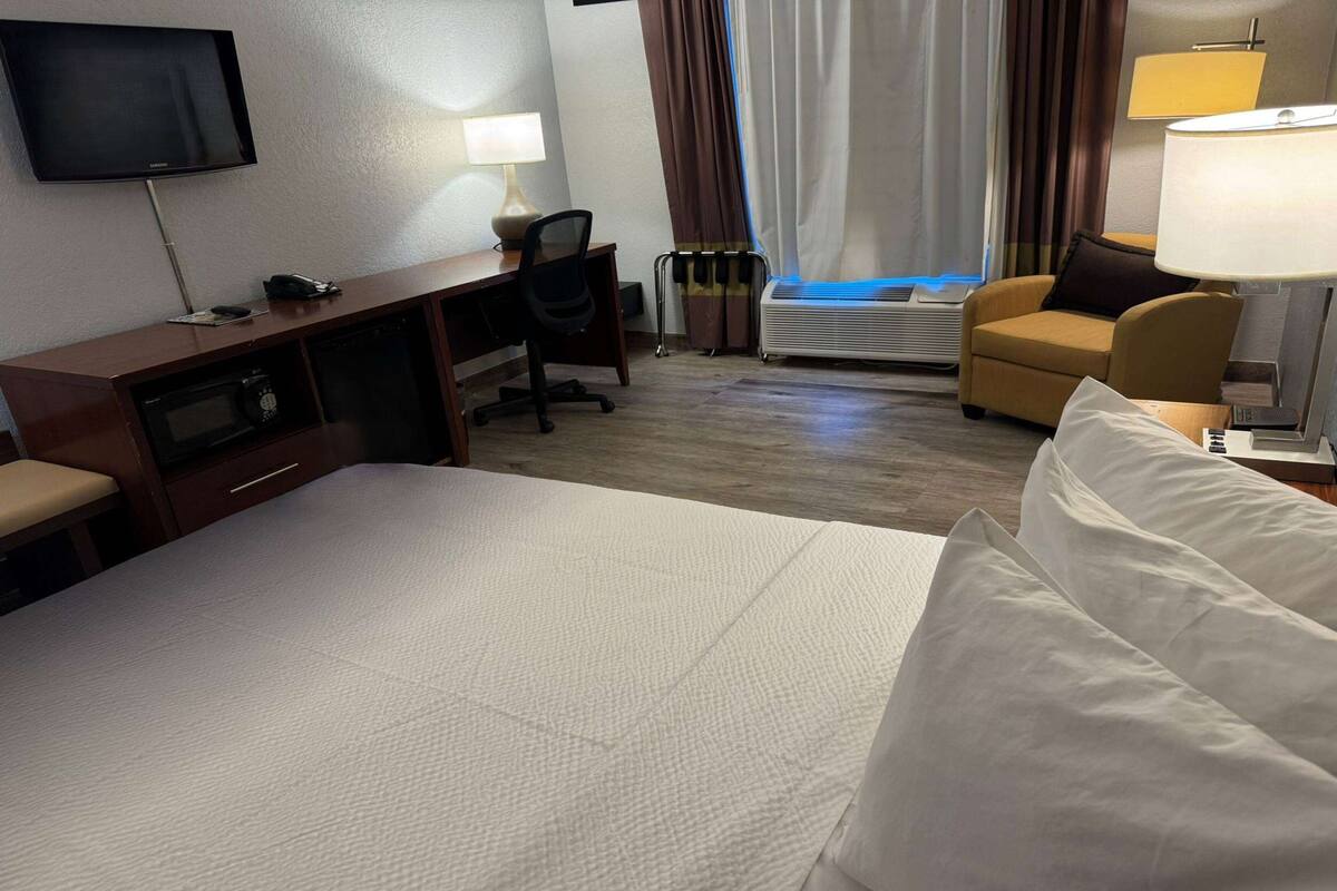 A hotel room with a bed, a desk with a chair, a television, an air conditioner, and a lamp.