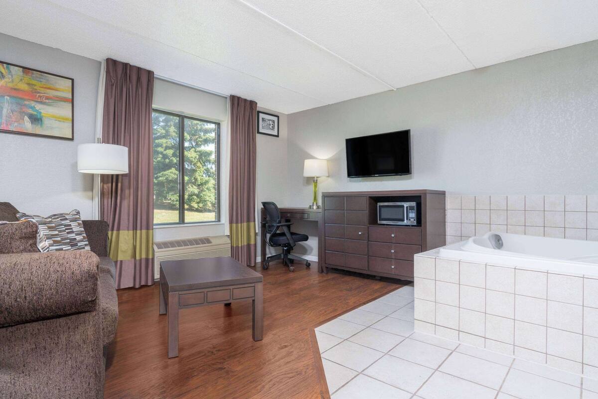 A hotel room with a sofa, a desk, a chair, a television, a microwave, and a bathtub.