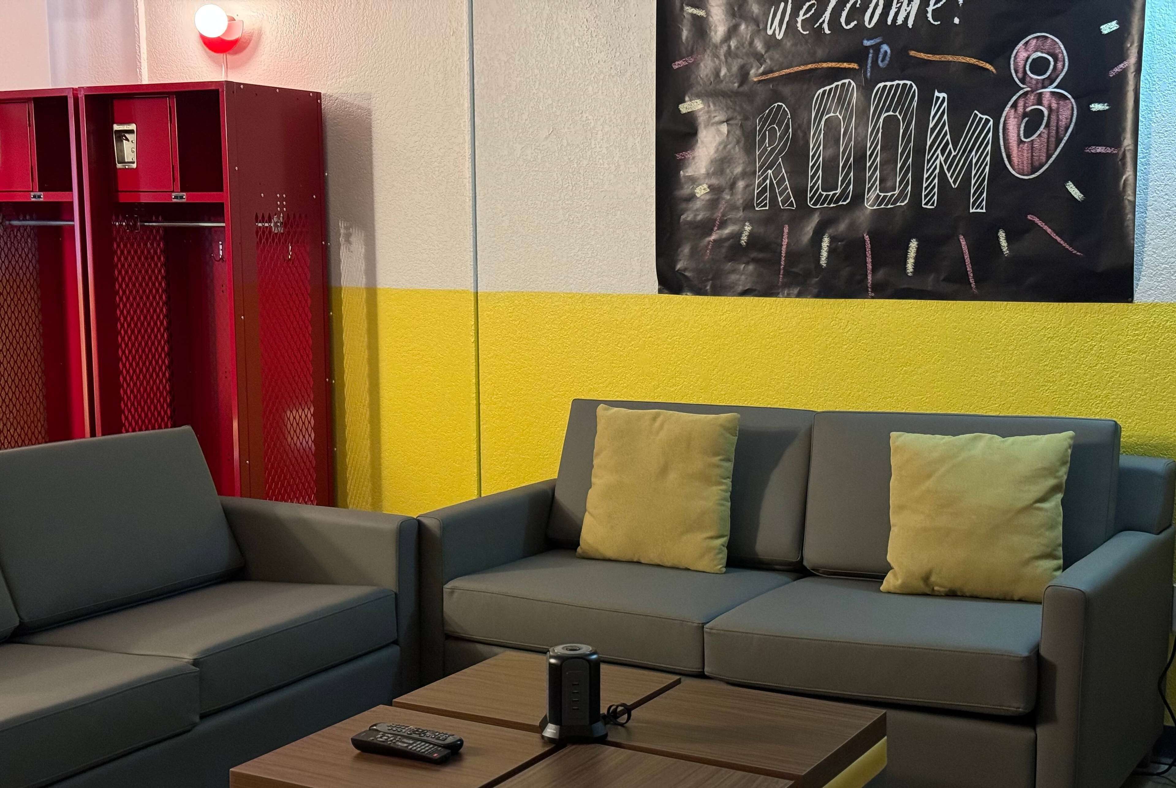 Une pièce avec un canapé gris, des coussins jaunes, une table basse en bois avec une télécommande, et un casier rouge.