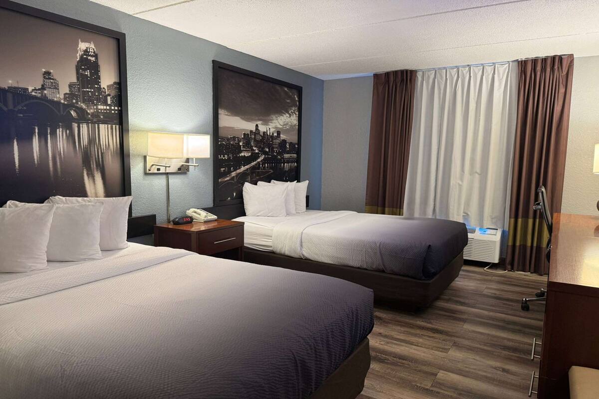 A hotel room with two beds, a nightstand with a phone, a wall-mounted lamp, and a large cityscape painting.
