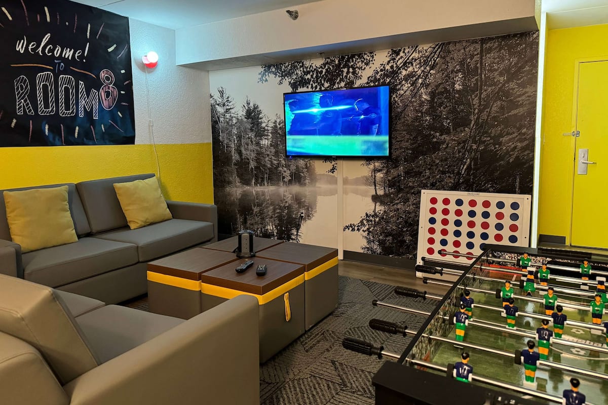 A modern living room with a sofa, a coffee table, a foosball table, a TV, and a wall with a welcome sign and a forest mural.