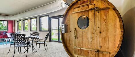 A sauna room with a wooden barrel door labeled "SAUNA," a seating area with metal chairs, and large windows allowing natural light.