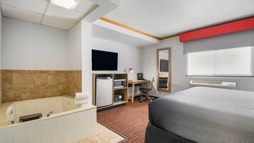 A hotel room with a large bed, a bathtub, a desk with a computer, a TV, and a microwave.