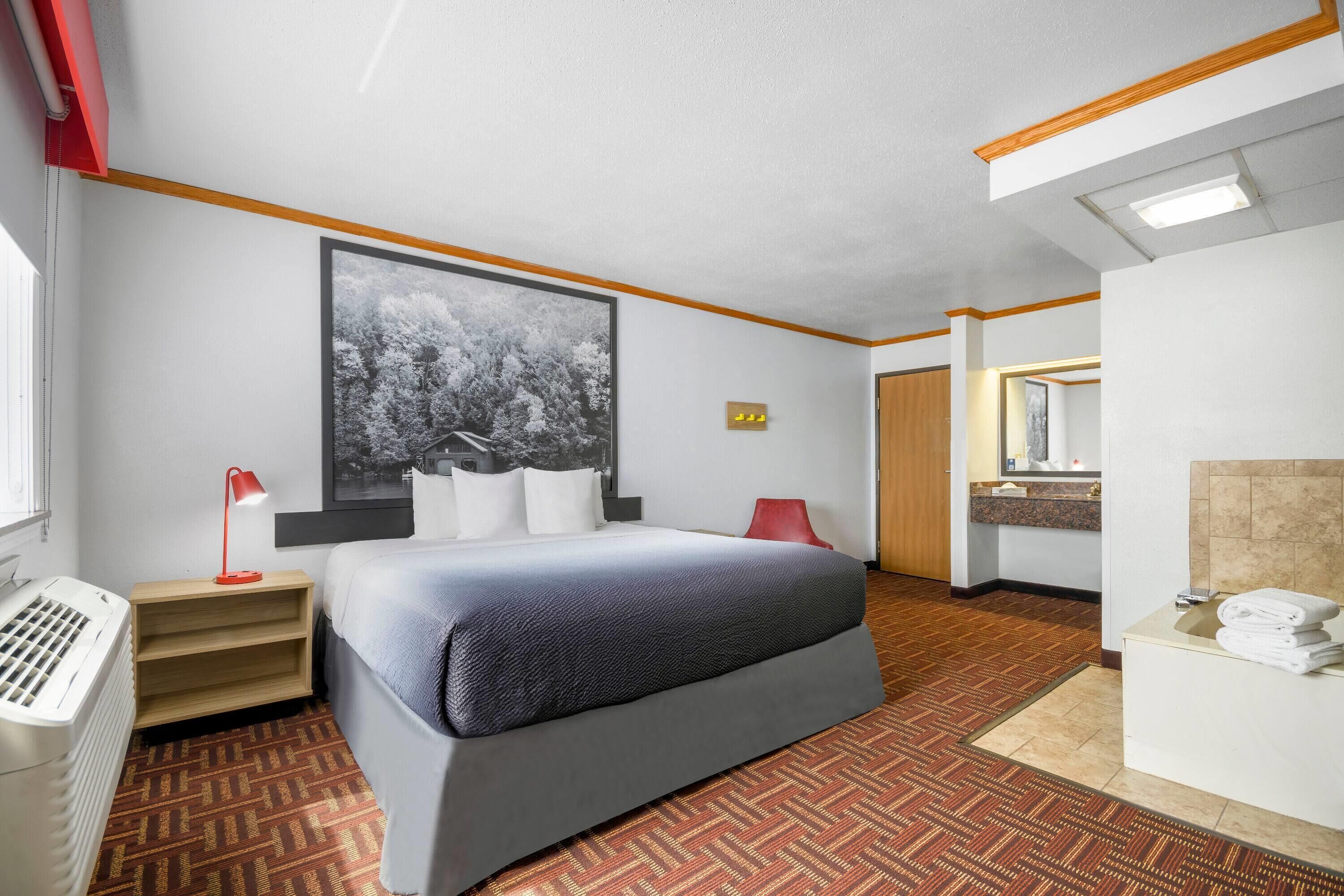 A hotel room with a large bed, a desk with a lamp, a red chair, a bathroom with a mirror, and a patterned carpet.