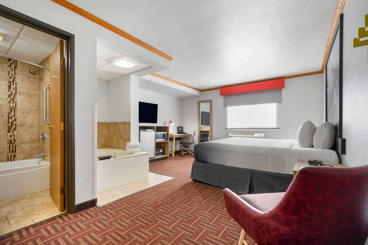 Une chambre d’hôtel avec un lit, un fauteuil rouge, un bureau avec un ordinateur, une télévision et une salle de bain avec une baignoire et une douche.