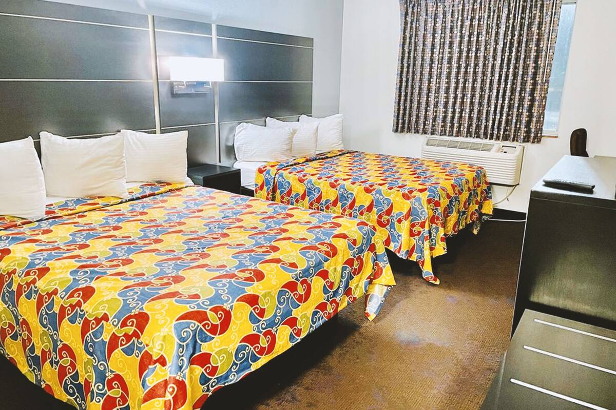 A hotel room with two beds, a headboard, a window with curtains, an air conditioning unit, and a desk with a lamp.