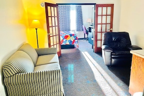 Quarto de hotel com sofá listrado, poltrona de couro preto, escrivaninha de madeira e cama com colcha colorida.