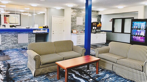 A hotel lobby with striped sofas, a wooden table, a kitchen area, and a refrigerator displaying 'Cold Drinks'. - Coratel Inn & Suites By Jasper Northfield (Northfield)