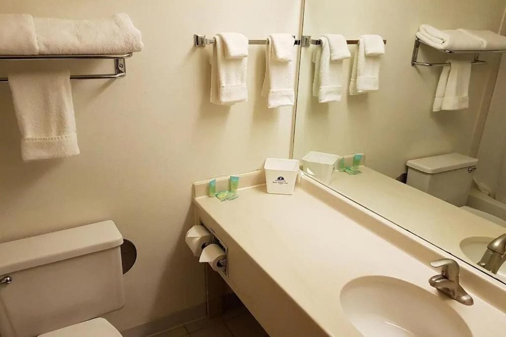 A bathroom with a toilet, sink, and mirror. There are towels hanging on a rack and a towel dispenser.