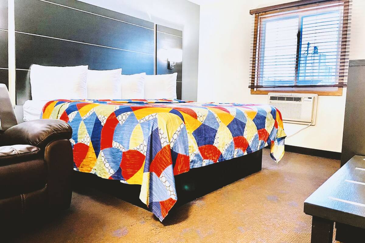 A hotel room with a large bed, a leather armchair, a small table, and a window with blinds.