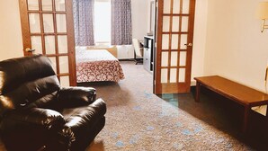 A hotel room with a leather sofa, a wooden bench, a bed with a floral bedspread, and a desk with a chair. - Coratel Inn & Suites By Jasper Northfield (Northfield)