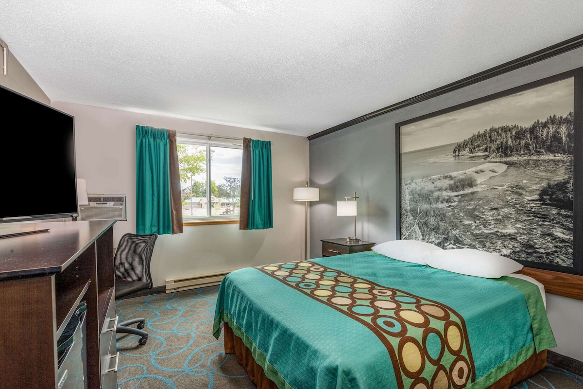A hotel room with a bed, a desk with a TV, a window with curtains, a lamp, and a painting on the wall.