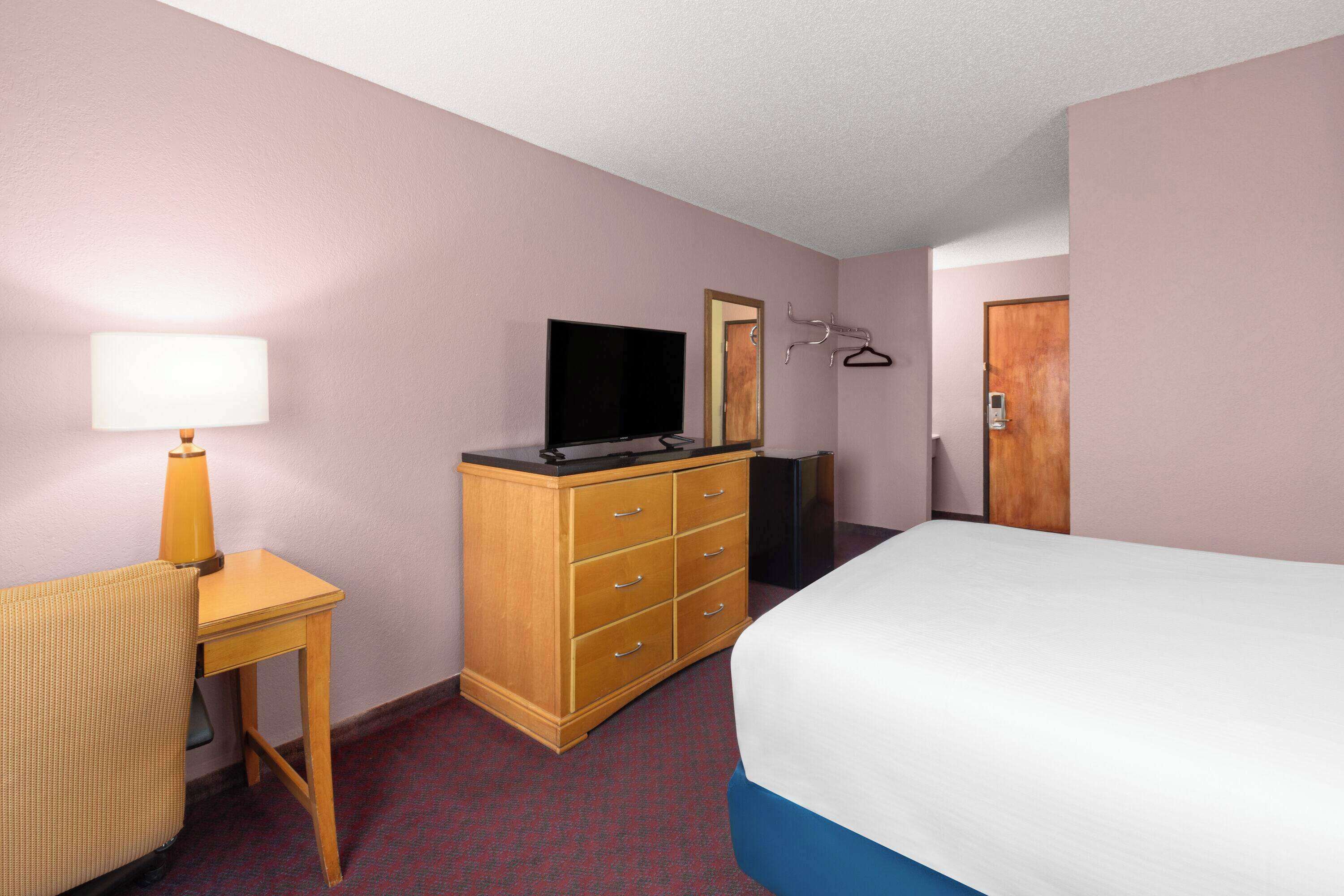 Habitación de hotel con cama, cómoda de madera, televisor, escritorio con lámpara y espejo.