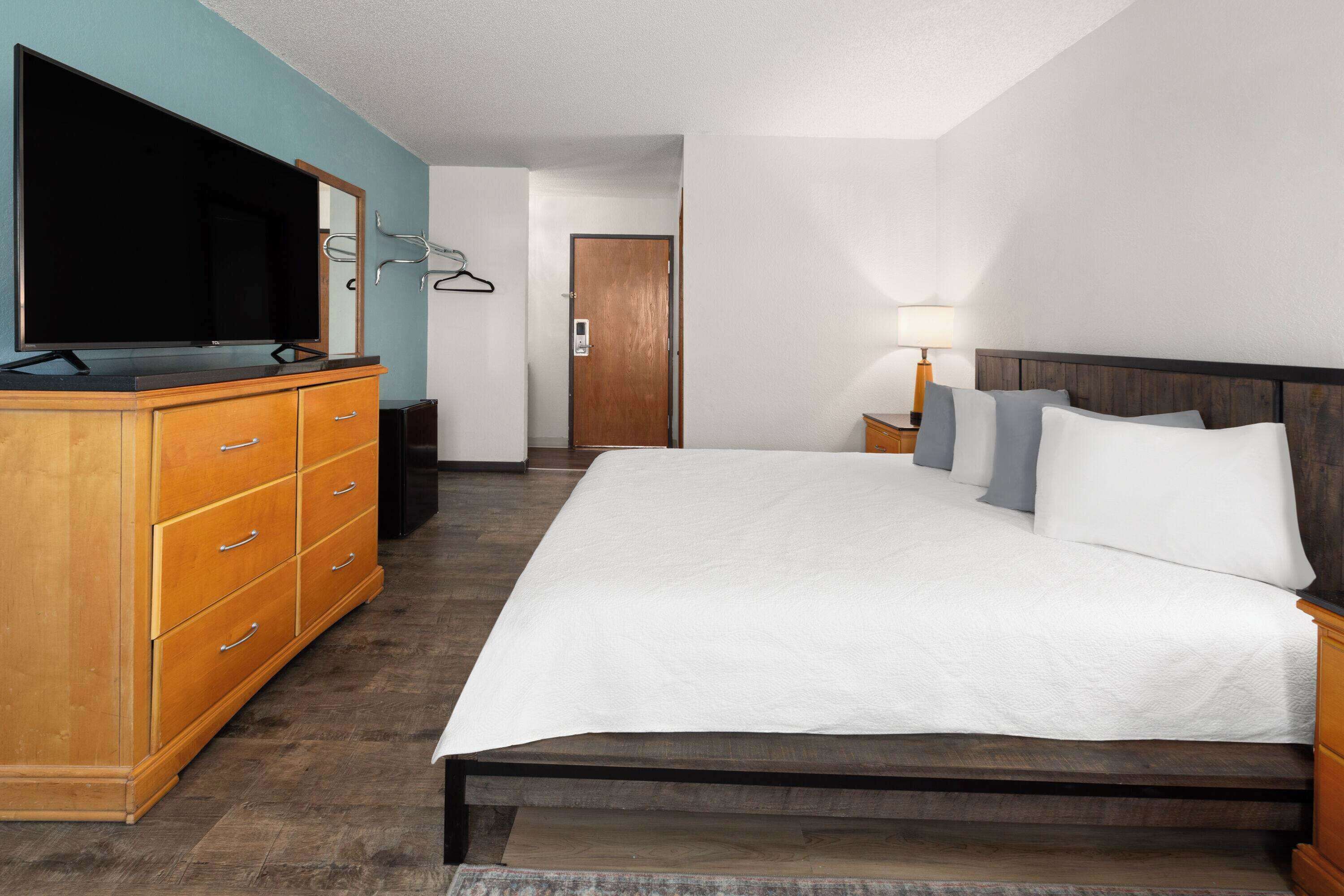 Quarto de hotel com uma cama grande, cómoda de madeira, televisão de ecrã plano e uma porta ao fundo.