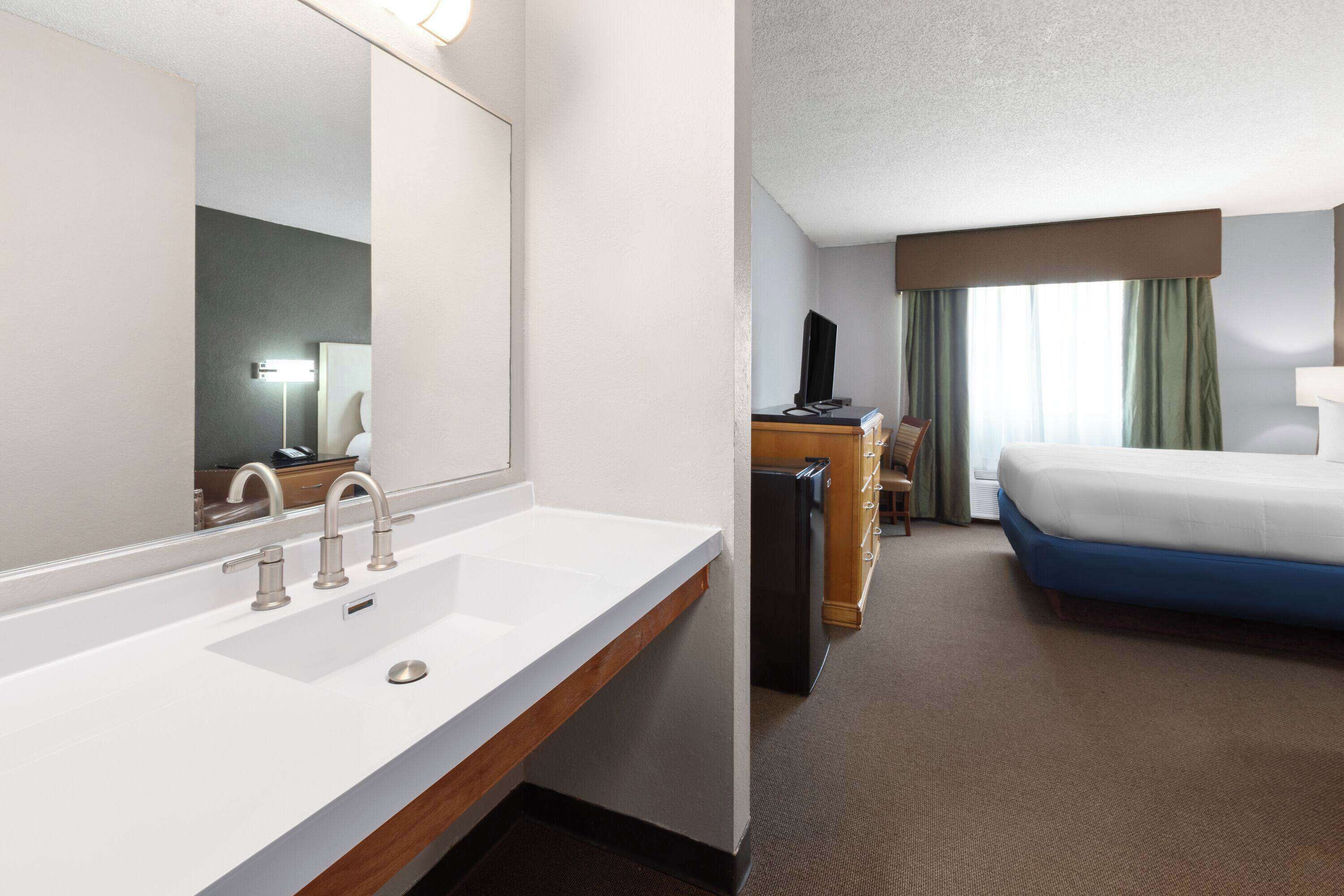 Une chambre d’hôtel avec un lavabo blanc, un miroir, un bureau avec un ordinateur et un lit avec un matelas bleu.