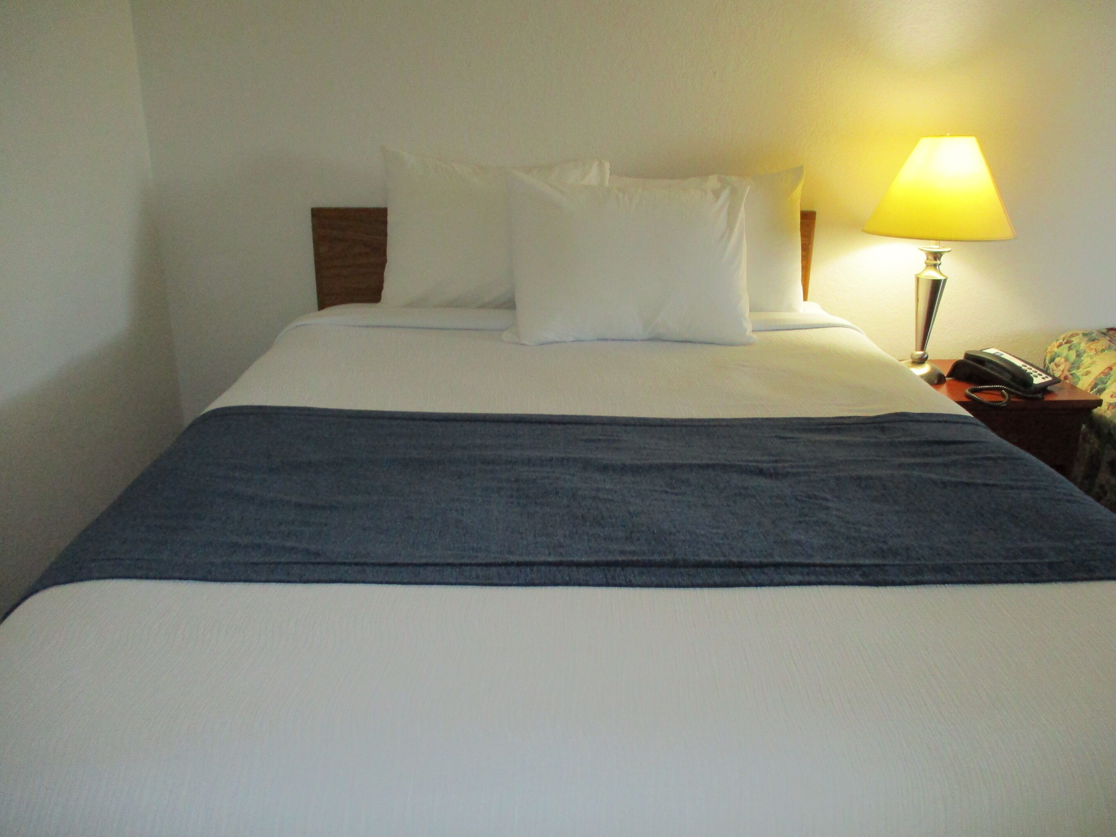 Una cama bien hecha con sábanas blancas y un plaid azul, una lámpara de noche y un teléfono sobre una mesita de madera.